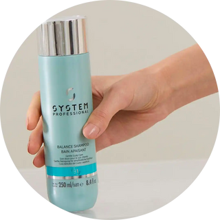 A hand holding a turquoise System Professional shampoo bottle.