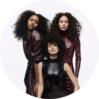 Three models with curly dark hair wearing sleek leather and vinyl outfits in burgundy and black tones.